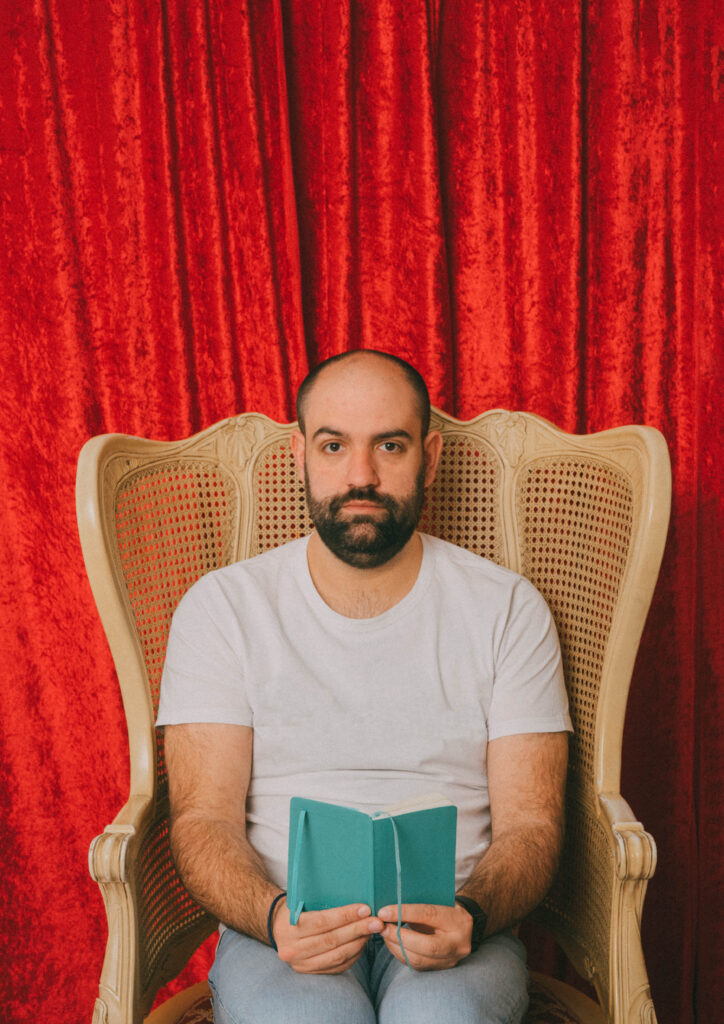 Hombre sentado en silla, cortina roja de fondo.