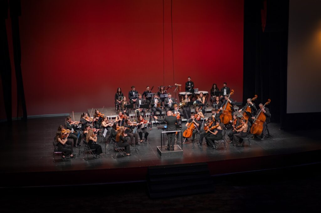 Orquesta sinfónica actuando en escenario con fondo rojo